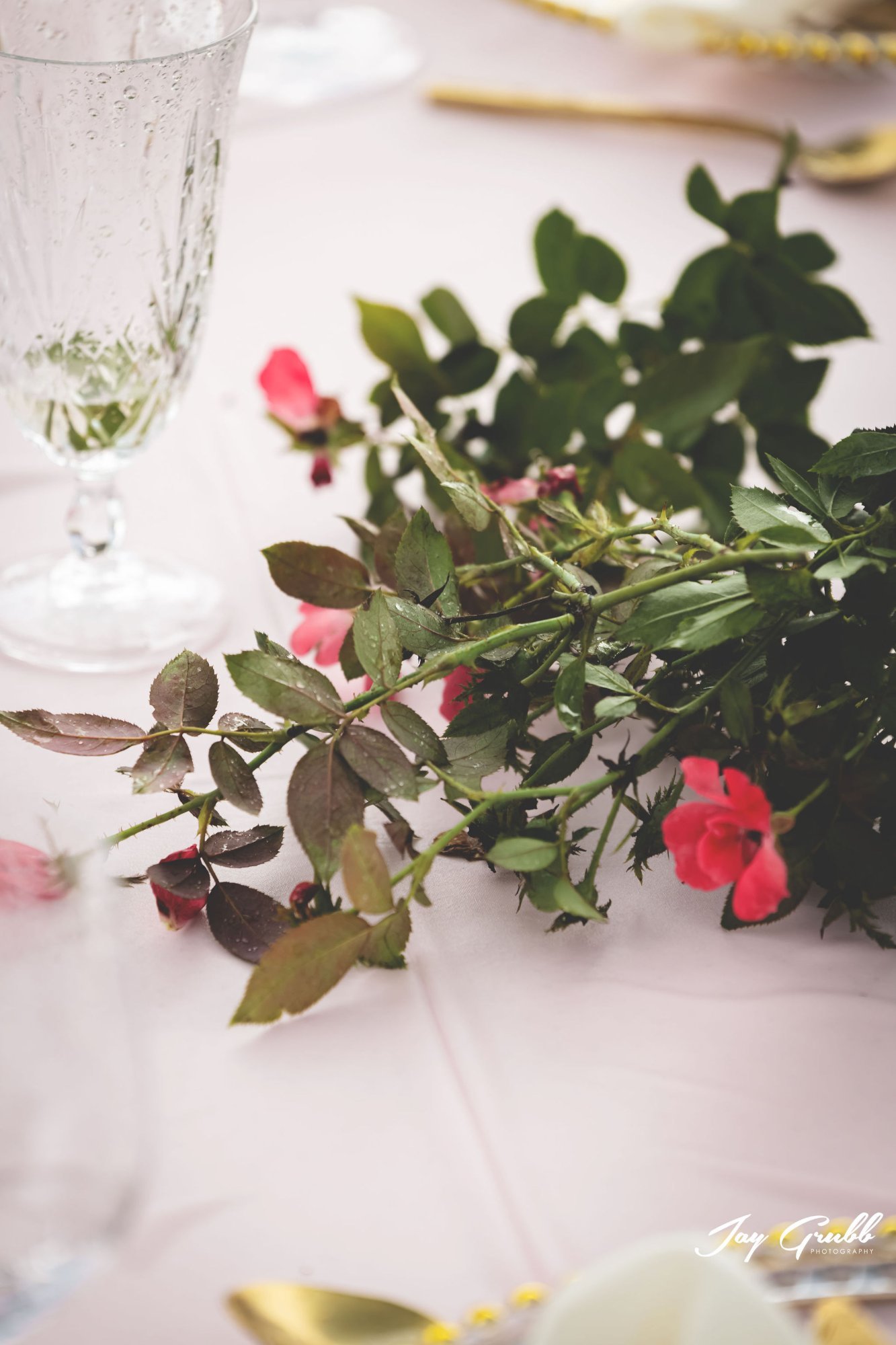 Pink garden roses laid on soft blush linen with crystal goblets and gold flatware