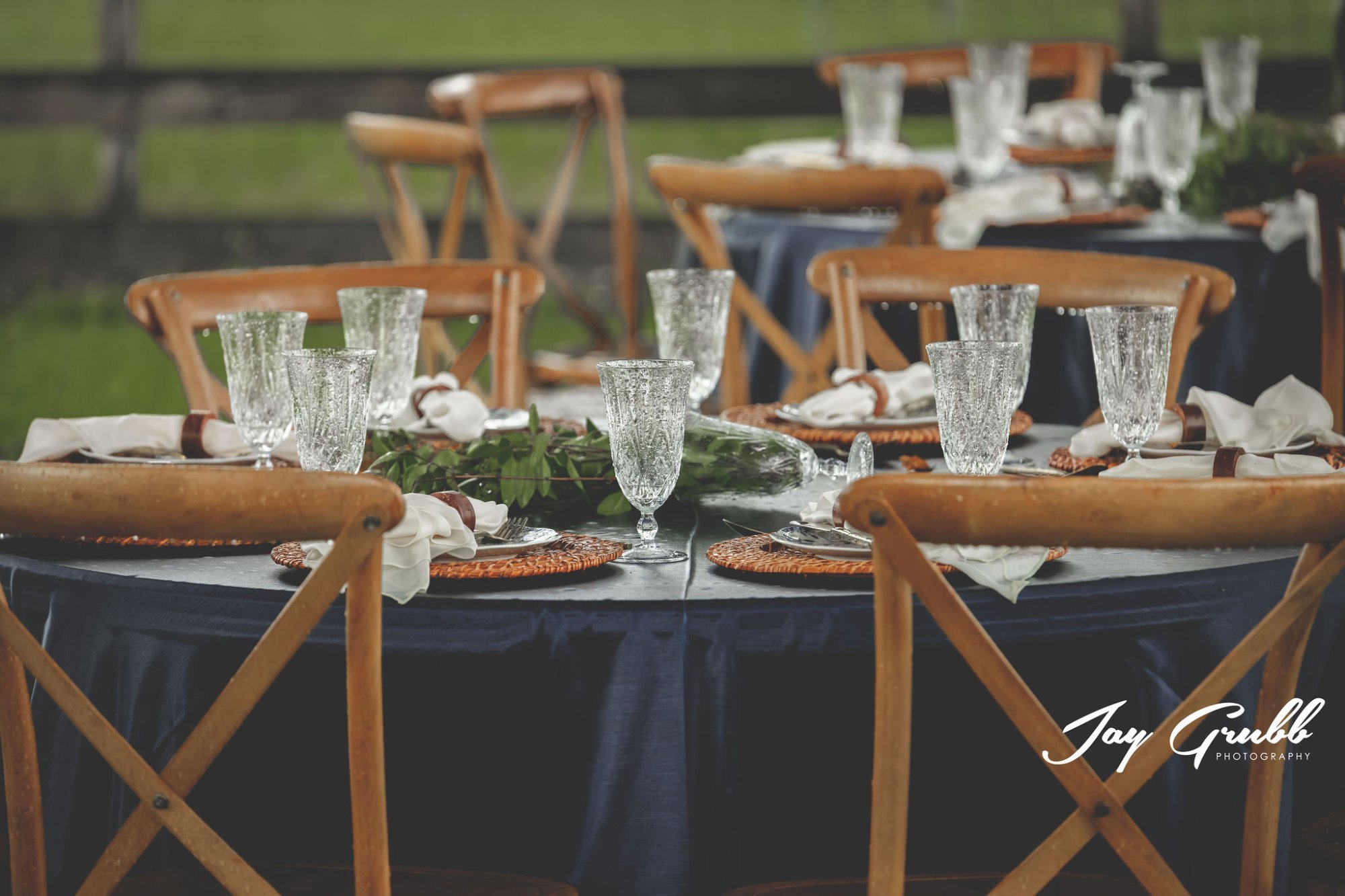 Reception tablescape with navy linens, cross-back chairs, wicker chargers, and a greenery runner
